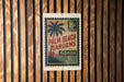 A vintage-style postage stamp with a palm tree design, hanging on a wooden wall.
