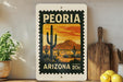 A vintage postage stamp from Arizona featuring a desert landscape with cacti and mountains, displayed on a shelf next to a wooden cutting board and some potted plants.
