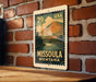 A metal sign with a vintage-style illustration of a mountain landscape and a river, mounted on a brick wall.