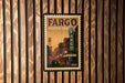 A framed poster of a city street with a car and a sign for the Fargo theater, mounted on a wooden wall.