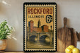 A framed vintage postage stamp depicting the Rockford, Illinois skyline, with a bridge and buildings, is displayed on a shelf next to a wooden cutting board and a potted plant.