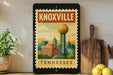 A framed poster of Knoxville, Tennessee, featuring a cityscape with a prominent tower, is displayed on a shelf next to a wooden cutting board and a potted plant.