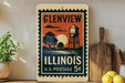 A vintage postage stamp from Illinois, featuring a water tower and a sunset landscape, is displayed on a shelf next to a wooden cutting board and some lemons.