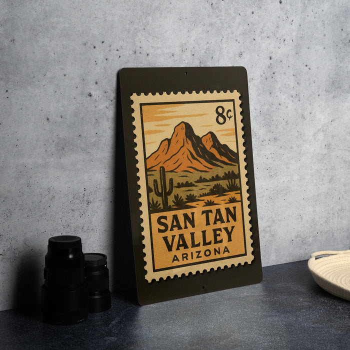 a metal sign with a desert landscape and mountains, along with a postage stamp design, placed on a countertop.
