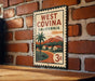 A vintage-style metal sign with the text "WEST COVINA CALIFORNIA" and a palm tree illustration, mounted on a brick wall.