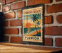 A vintage-style metal stamp depicting a tropical beach scene with palm trees and a sunset, mounted on a brick wall.