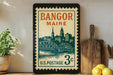 A framed vintage postage stamp from Bangor, Maine, featuring a cityscape and a bridge, is displayed on a shelf next to a wooden cutting board and some lemons.