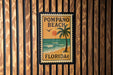 A vintage-style stamp or sign for Pompano Beach, Florida, featuring a palm tree and a sunset over the ocean.