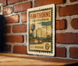 A vintage-style postage stamp from the United States, featuring a scene of a town and palm trees, mounted on a brick wall.