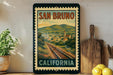 A vintage-style poster of San Bruno, California, featuring a train track and a town in the background, is displayed on a wall next to a wooden cutting board and some potted plants.