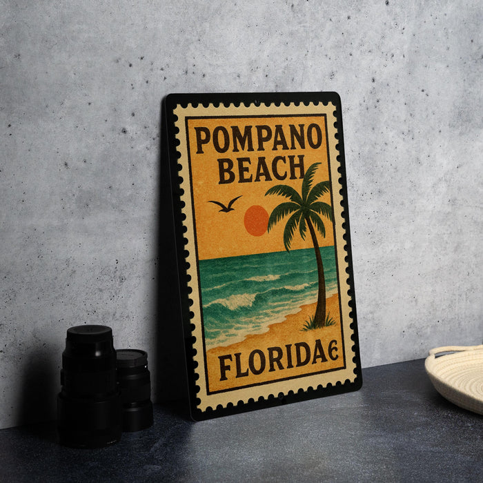 A framed poster of Pompano Beach, Florida, featuring a palm tree and ocean scene, is displayed on a countertop next to some black camera lenses.