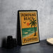 A framed poster of Pompano Beach, Florida, featuring a palm tree and ocean scene, is displayed on a countertop next to some black camera lenses.