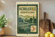 A vintage-style postage stamp from Kentucky, featuring a pastoral scene with a church and a farm, is displayed on a shelf next to kitchen utensils and a potted plant.