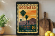 A vintage-style postage stamp from California, featuring a palm tree and mountains, is displayed on a shelf next to a wooden cutting board and a potted plant.