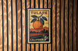 A vintage postage stamp from Tulare, California, featuring an orange and a leaf, is displayed on a wooden wall.