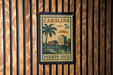 A vintage-style postage stamp from Carolina, featuring a palm tree and buildings, is displayed on a wooden wall.