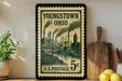 A vintage U.S. postage stamp from Youngstown, Ohio, featuring steam-powered factories and a bridge, displayed on a shelf next to a wooden cutting board and some lemons.