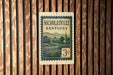 A vintage-style postage stamp from Kentucky, featuring a church and a rural landscape, is displayed on a wooden wall.