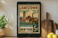 A framed vintage postage stamp depicting a cityscape of Jamestown, New York, with the text "JAMESTOWN NEW YORK 5c" printed on it. The stamp is displayed on a shelf next to a wooden cutting board and a potted plant.