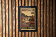 A framed poster of the scenic falls in South Dakota, featuring a waterfall and a building, is displayed on a wooden wall.
