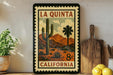 A framed vintage-style stamp depicting a desert landscape with a cactus and mountains, along with the text "LA QUINTA CALIFORNIA 8¢".