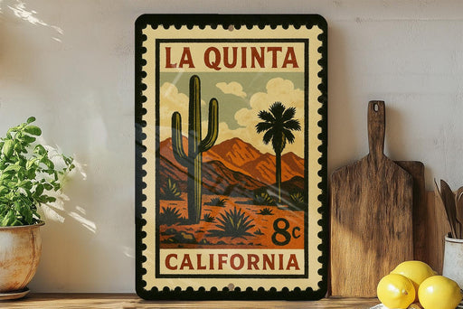 A framed vintage-style stamp depicting a desert landscape with a cactus and mountains, along with the text "LA QUINTA CALIFORNIA 8¢".