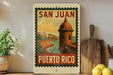 A vintage-style poster of San Juan, Puerto Rico, featuring a castle and a coastal landscape, is displayed on a wall next to a wooden cutting board and a potted plant.