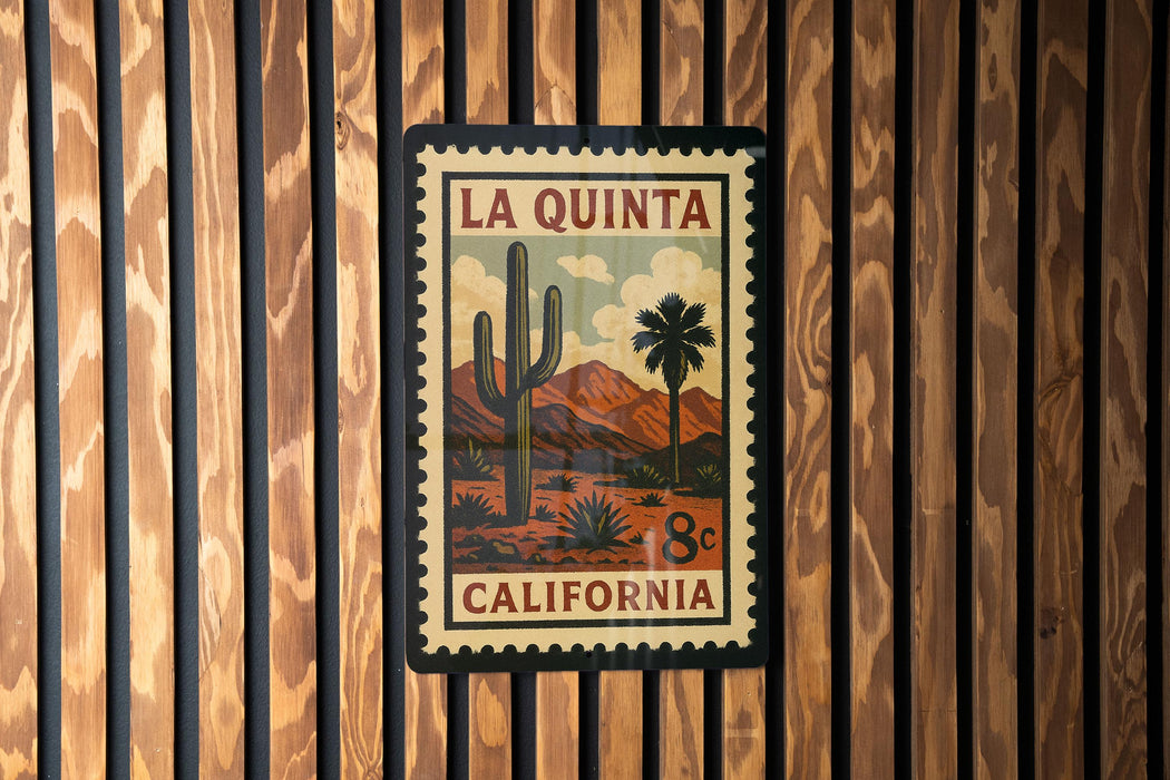 A framed vintage-style stamp depicting a desert landscape with a cactus and mountains, mounted on a wooden wall.