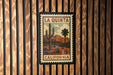 A framed vintage-style stamp depicting a desert landscape with a cactus and mountains, mounted on a wooden wall.