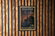 A framed Massachusetts postage stamp depicting a house and a church, mounted on a wooden wall.