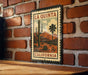 A metal sign with a vintage-style illustration of a desert landscape, featuring a cactus and mountains, mounted on a brick wall.