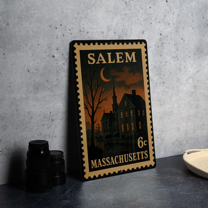 A framed vintage postage stamp from Salem, Massachusetts, depicting a house and a church at night, placed on a countertop next to some black camera lenses.
