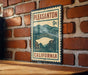 A vintage-style metal sign with the text "PLEASANTON 9c" and "CALIFORNIA" printed on it, mounted on a brick wall.