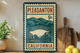 A vintage-style metal sign with the text "PLEASANTON 9c" and an illustration of a small town, with the text "CALIFORNIA" below it. The sign is displayed on a shelf next to a wooden cutting board and a potted plant.