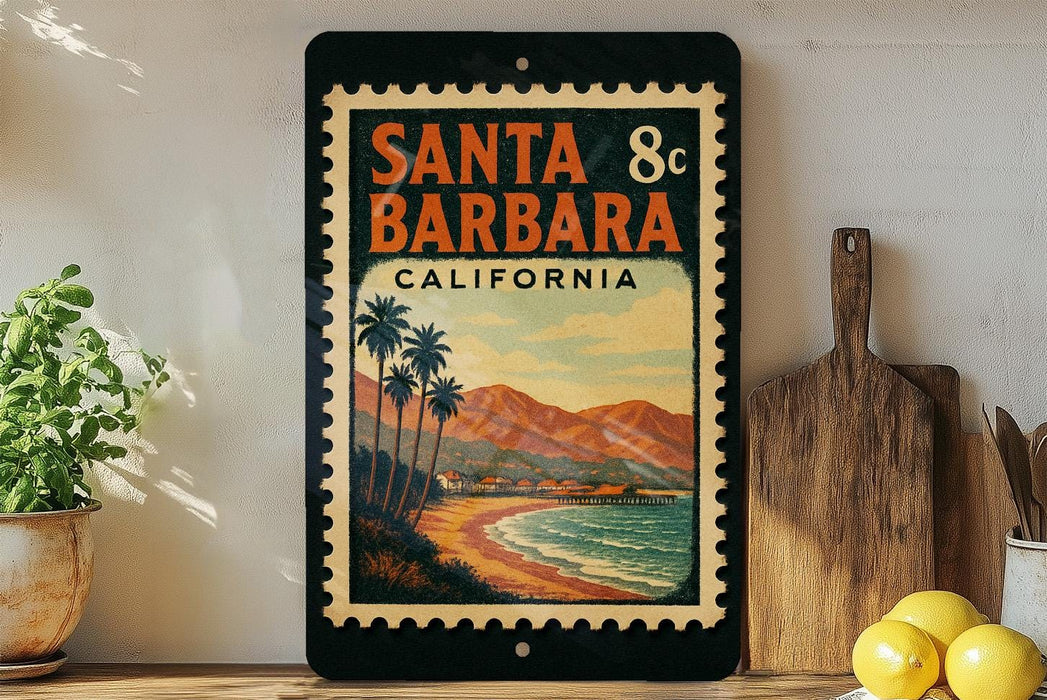A vintage-style metal sign with a beach scene and mountains in the background, featuring the text "SANTA BARBARA, CALIFORNIA" and a postage stamp-like border.
