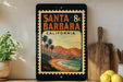 A vintage-style metal sign with a beach scene and mountains in the background, featuring the text "SANTA BARBARA, CALIFORNIA" and a postage stamp-like border.