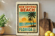 A vintage-style postage stamp depicting a beach scene with palm trees and a sunset, placed on a shelf next to a wooden cutting board and some lemons.