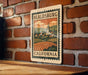 A vintage-style metal sign with a stamp-like border hangs on a brick wall, depicting a rural landscape with a church steeple and a farm in the foreground.