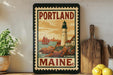 A framed vintage postage stamp from Maine, featuring a lighthouse and a sailboat, is displayed on a shelf next to a wooden cutting board and a potted plant.