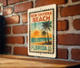 A vintage-style metal sign with a tropical beach scene and palm trees, mounted on a brick wall.