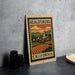 A metal sign with a vintage-style illustration of a town and countryside, featuring a church and a mountain in the background, is displayed on a table.