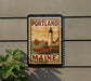 A vintage-style postage stamp depicting a lighthouse and sailboat, with the text "PORTLAND MAINE" and "U.S. POSTAGE" printed on it, is displayed on a wall.