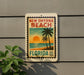 A vintage-style postage stamp depicting a tropical beach scene with palm trees and a sunset, mounted on a wall.