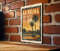 A vintage-style metal sign with the text "LA PALMA CALIFORNIA" displayed on it, featuring a palm tree and a building in the background.