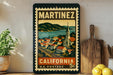 A vintage-style postage stamp from California, featuring a colorful illustration of a town and mountains, displayed on a shelf next to a wooden cutting board and some potted plants.
