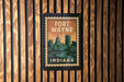 A vintage-style metal sign with the text "FORT WAYNE INDIANA" displayed on it, mounted on a wooden wall.
