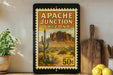 A framed vintage postage stamp depicting the Apache Junction in Arizona, with a desert landscape and cacti, is displayed on a shelf next to a wooden cutting board and a potted plant.