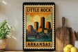 A framed vintage-style poster of Little Rock, Arkansas, featuring a cityscape with a bridge and a sun in the sky.