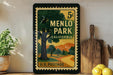 A vintage-style postage stamp from Menlo Park, California, featuring a scenic landscape with a winding road and trees. The stamp is displayed on a shelf next to a wooden cutting board and a potted plant.