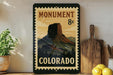 A vintage-style metal sign with a mountain landscape and the text "MONUMENT COLORADO" is displayed on a shelf, accompanied by a wooden cutting board and a potted plant.